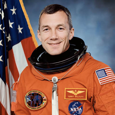 Headshot of astronaut Terry Wilcott in orange space suit minus helmet in fron the of the U.S. flag