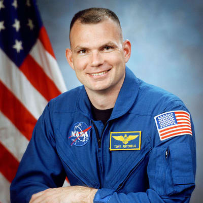 Headshot of astronaut Tony Antonelli in blue flight suit in front of the U.S. fla