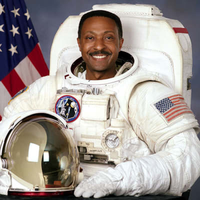 Headshot of astronaut Winston Scott in full space suit holding his helmet and standing in front of the U.S. flag