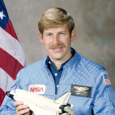 Headshot of astronaut Woody Spring in blue flight suit holding a model of the space shuttle in front of the U.S. flag