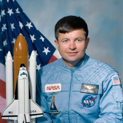 Headshot of astronaut Ken Cameron in blue flight suit in front of the U.S. flag and next to a model of the space shuttl