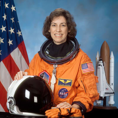 Headshot of Ellen Ochoa in an orange flight suit, holding her flight helmet and gloves. In the background is the U.S. flag and a model of the space shuttle.
