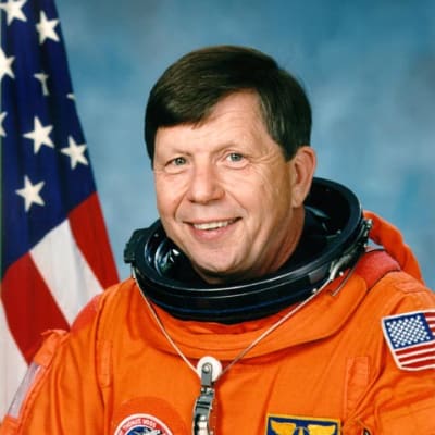 Headshot of astronaut Roger Crouch in orange space suit without helmet in front of a U.S. flag