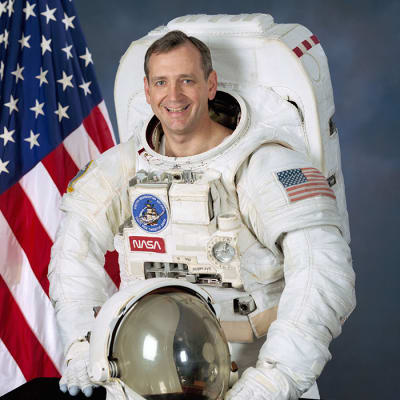 Official NASA headshot of astronaut Tom Akers, wearing a space suit with the American flag in the background