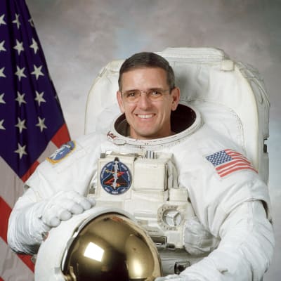 Headshot of astronaut Bill McArthur in space suit holding his helmet in front of U.S. flag