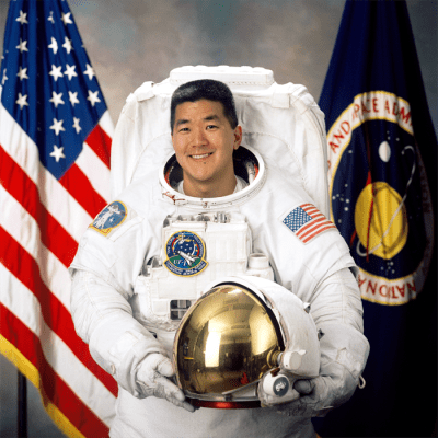 Astronaut Daniel Tani in white flight suit holding his helmet in front of the American flag and NASA flag