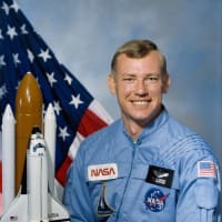 Fotografía del astronauta Mark Brown con traje de vuelo azul delante de la bandera de EE.UU. y junto a una maqueta del transbordador espacial.
