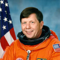 Headshot of astronaut Roger Crouch in orange space suit without helmet in front of a U.S. flag
