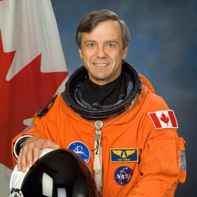 Astronaut Robert “Bob” Thirsk wearing an orange flight suit and standing in front of a Canadian flag