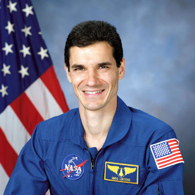 Headshot photo of NASA astronaut Gregory Linteris wearing a blue flight suit in front of the American flag