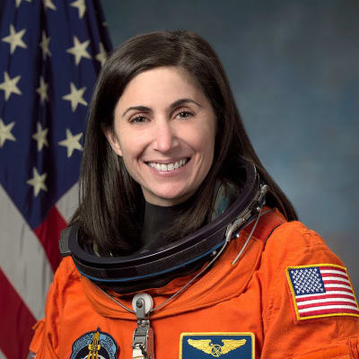 Headshot of astronaut Nicole Stott in orange space suit without helmet in front of the U.S. flag