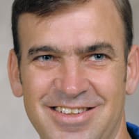 Headshot of astronaut John Fabian in blue flight suit in front of a space shuttle model and flag with official NASA seal
