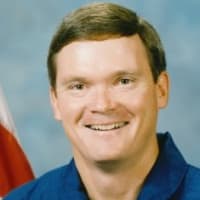 Astronaut Bruce Melnick in blue flight suit standing with arms crossed in front of space shuttle Atlantis