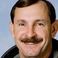 Headshot of astronaut Curt Brown in orange space suit holding his helmet in front of a U.S. flag and model of the space shuttle