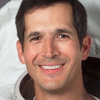 Headshot of astronaut Commander John Herrington in full space suit, holding his helmet in front of the U.S. and NASA flags