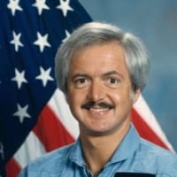 Astronaut John Bartoe in blue flight suit in front of the American flag and next to a model of the space shuttle