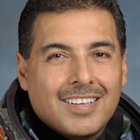 Headshot of astronaut José Hernandez in orange space suit, holding his helmet and standing in front of the U.S. flag and a model of the space shuttle