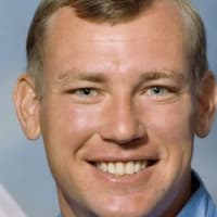Headshot of astronaut Mark Brown in blue flight suit in front of the U.S. flag and next to a model of the space shuttle