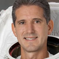Headshot of astronaut Mike Good in full space suit holding helmet in front of the U.S. flag