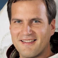 Astronaut Richard Hieb in full space suit holding his helmet in front of U.S. flag