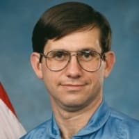 Photo of NASA astronaut Robert Cenker next to the United States flag and a model of the space shuttle