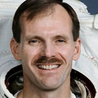 Astronaut Steven Smith in full space suit holding his helmet in front of the U.S. flag