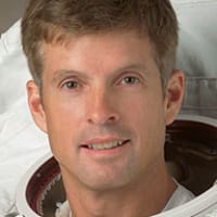 Headshot of astronaut Steve Swanson in full space suit holding his helmet in front of the U.S. flag