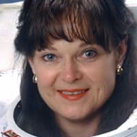 Headshot of astronaut Tammy Jernigan in full space suit holding her helmet in front of the NASA flag and U.S. flag