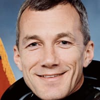 Headshot of astronaut Terry Wilcott in orange space suit minus helmet in fron the of the U.S. flag