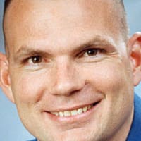 Headshot of astronaut Tony Antonelli in blue flight suit in front of the U.S. fla
