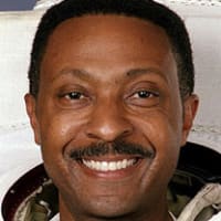 Headshot of astronaut Winston Scott in full space suit holding his helmet and standing in front of the U.S. flag