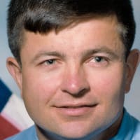Headshot of astronaut Ken Cameron in blue flight suit in front of the U.S. flag and next to a model of the space shuttl