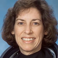 Headshot of Ellen Ochoa in an orange flight suit, holding her flight helmet and gloves. In the background is the U.S. flag and a model of the space shuttle.