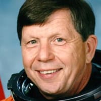 Headshot of astronaut Roger Crouch in orange space suit without helmet in front of a U.S. flag