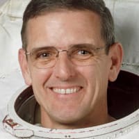 Headshot of astronaut Bill McArthur in space suit holding his helmet in front of U.S. flag