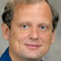Headshot of astronaut Norman Thagard in blue flight suit in front of NASA flag and space shuttle model