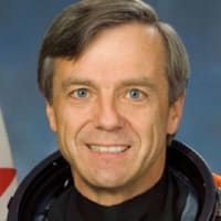 Astronaut Robert “Bob” Thirsk wearing an orange flight suit and standing in front of a Canadian flag