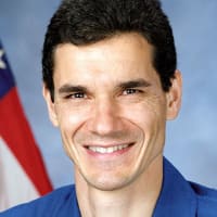 Headshot photo of NASA astronaut Gregory Linteris wearing a blue flight suit in front of the American flag