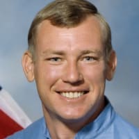 Headshot of astronaut Mark Brown in blue flight suit in front of the U.S. flag and next to a model of the space shuttle