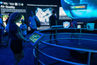 A blonde woman in shorts touches a monitor attached to a circular railing that surrounds a floating Earth. On the Earth, heat data is projected.