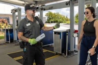 Security guard holding a metal detecting wand motioning for a female guest to walk through the metal detector.