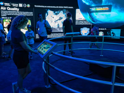 A blonde woman in shorts touches a monitor attached to a circular railing that surrounds a floating Earth. On the Earth, heat data is projected.