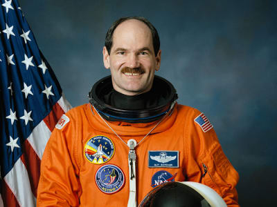 Headshot of astronaut Guy Gardner in orange flight suit holding helmet