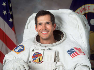Headshot of astronaut Commander John Herrington in full space suit, holding his helmet in front of the U.S. and NASA flags