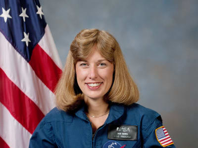 Headshot of astronaut Jan Davis in blue flight suit holding a model space shuttle in front of the U.S. flag