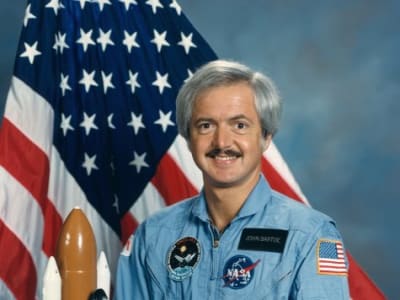 Astronaut John Bartoe in blue flight suit in front of the American flag and next to a model of the space shuttle