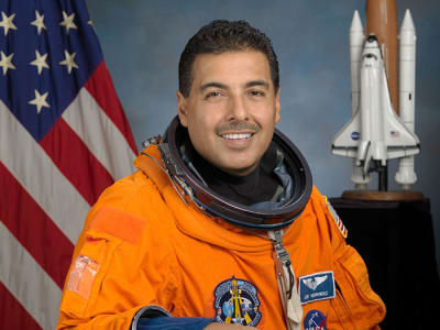 Headshot of astronaut José Hernandez in orange space suit, holding his helmet and standing in front of the U.S. flag and a model of the space shuttle