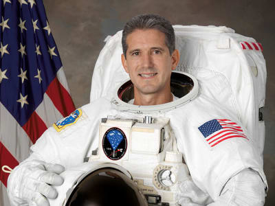 Headshot of astronaut Mike Good in full space suit holding helmet in front of the U.S. flag