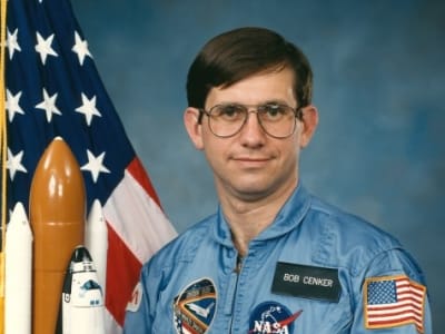 Photo of NASA astronaut Robert Cenker next to the United States flag and a model of the space shuttle