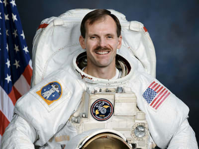 Astronaut Steven Smith in full space suit holding his helmet in front of the U.S. flag